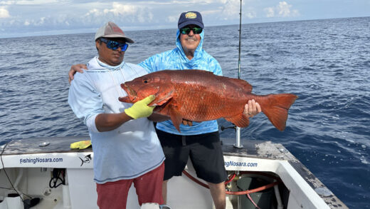 Two anglers on a boat, one holding a large red snapper they've just caught, with the ocean and a clear sky in the background.